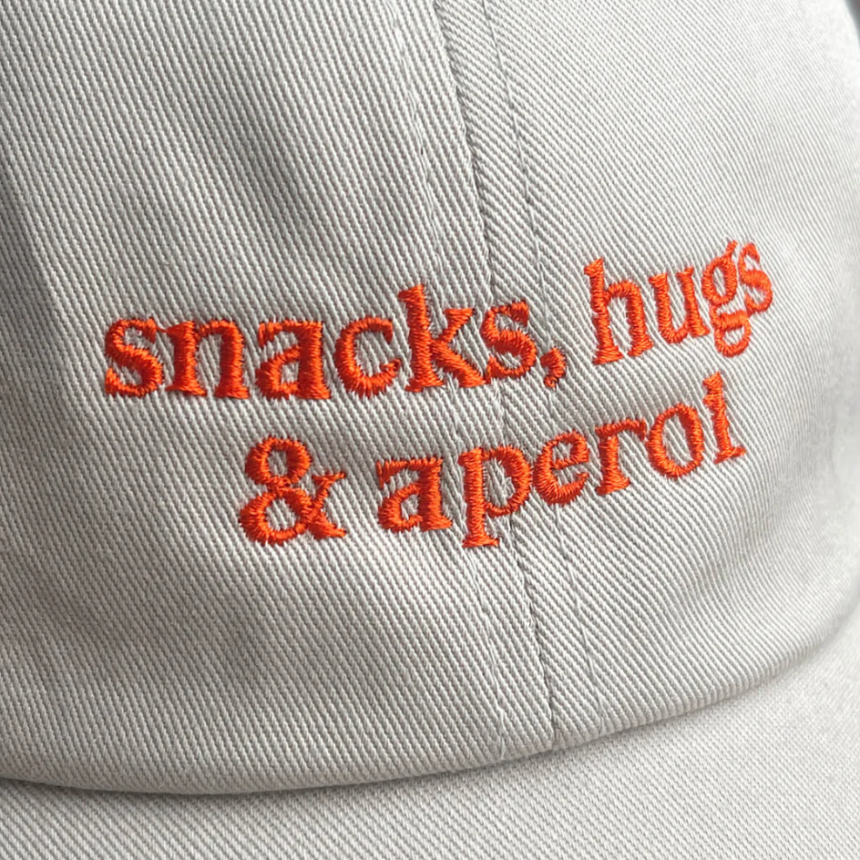 Cap mit gebogenem Schirm und Schriftzug „snacks, hugs & aperol“ in orange auf beigem Baumwollstoff, Studio Ciao