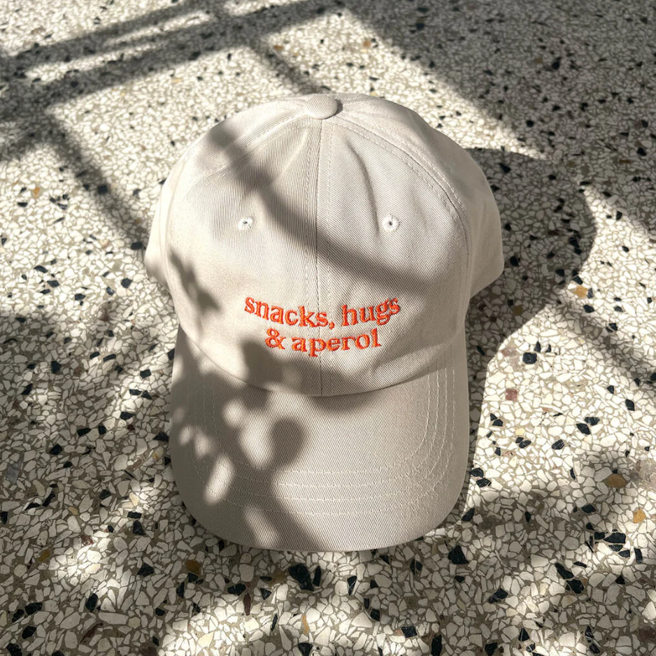 Cap mit gebogenem Schirm und Schriftzug „snacks, hugs & aperol“ in orange auf beigem Baumwollstoff, Studio Ciao